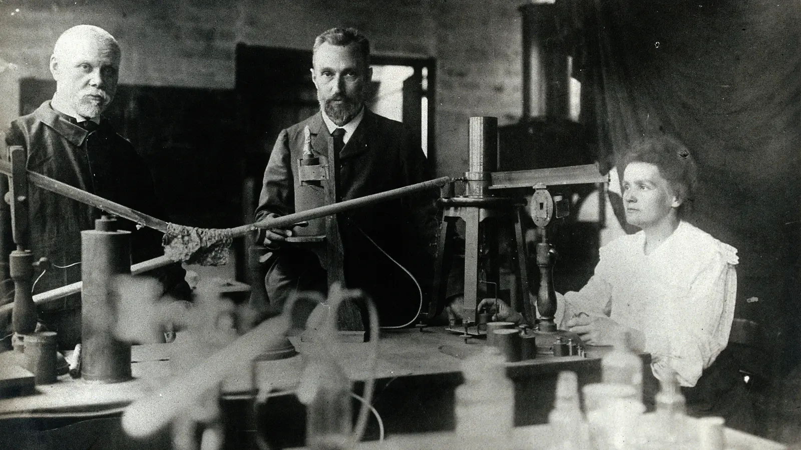 Pierre et Marie Curie dans leur laboratoire, debout et assise devant leurs instruments, avec un collègue à gauche