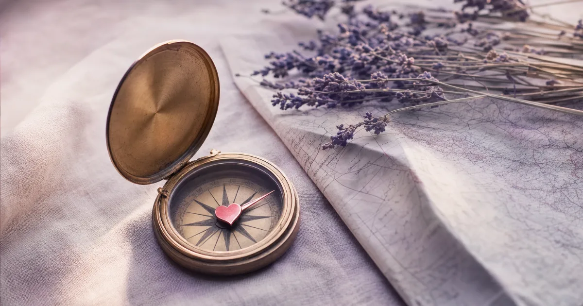 Une boussole en laiton ouverte sur une nappe en lin, l'aiguille remplacée par un petit cœur rose, à côté d'une carte pliée et d'un brin de lavande