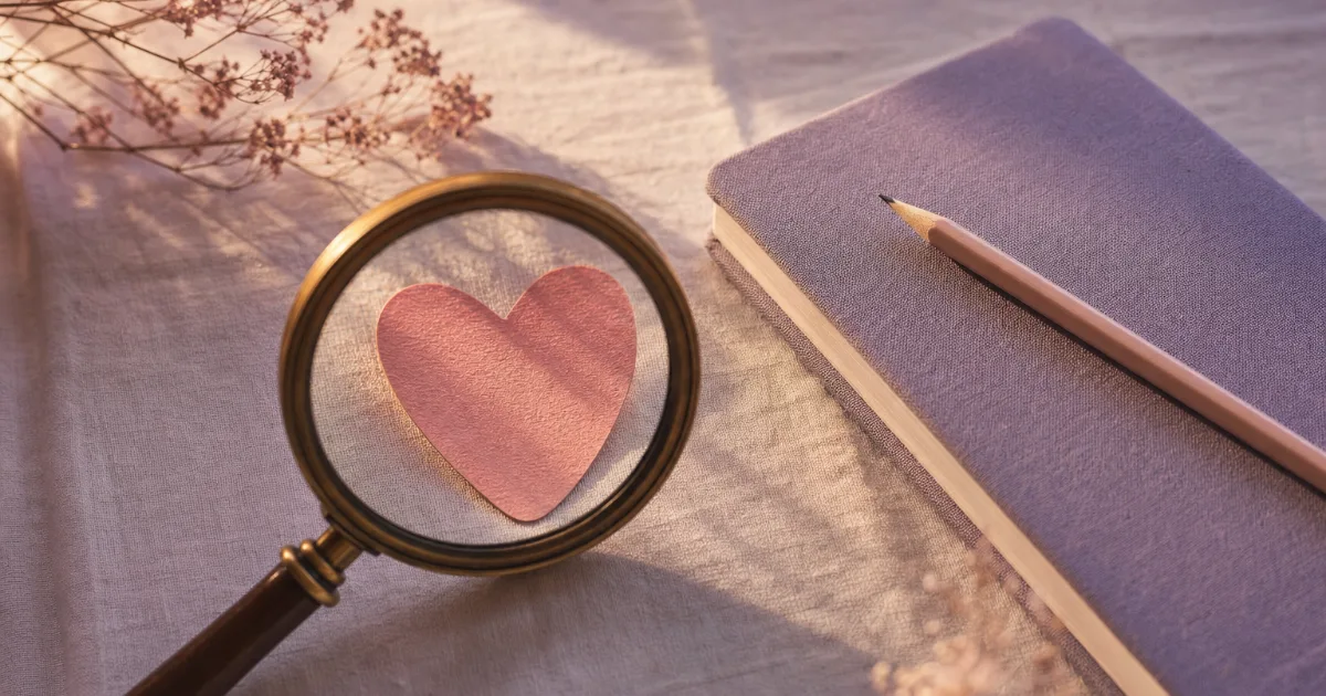 Une loupe ancienne à manche en bois posée sur une nappe en lin, sa lentille au-dessus d'un petit cœur en papier rose, à côté d'un carnet lavande fermé et d'un crayon