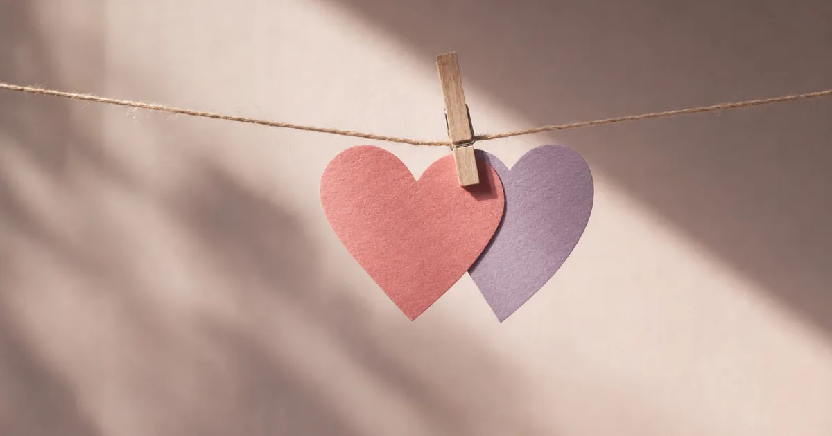 Deux cœurs en papier, un rose et un lavande, accrochés ensemble par une petite pince en bois sur une ficelle tendue