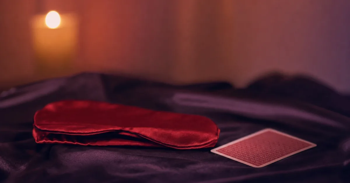 Un bandeau de soie rouge plié et une carte à jouer face cachée sur des draps de satin sombre, une bougie floue en arrière-plan